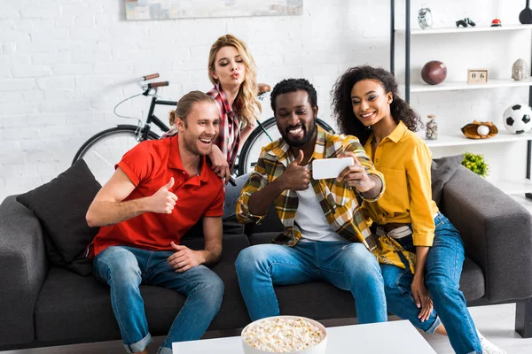 Excited happy multicultural men and women sitting on couch and taking selfie in living room — Stock Photo