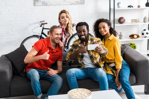 Animados homens e mulheres multiculturais felizes sentados no sofá e tomando selfie na sala de estar — Fotografia de Stock