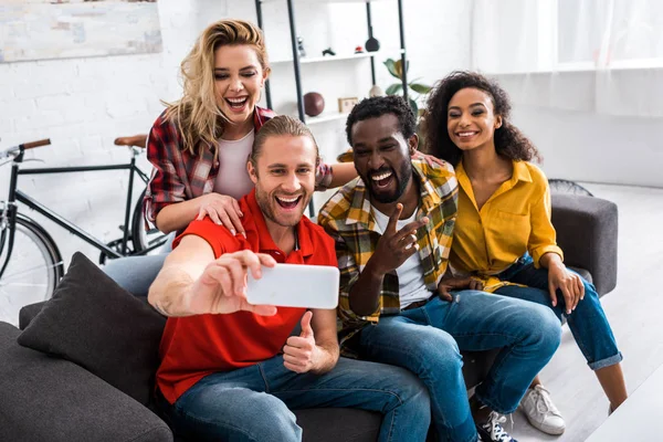 Foco seletivo de amigos multiculturais sorridentes sentados no sofá e tomando selfie em casa — Fotografia de Stock