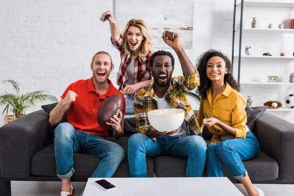 Happy multiethnic friends sitting on couch, watching match and holding bowl of popcorn — Stock Photo