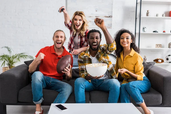 Amigos multiétnicos felizes sentados no sofá, assistindo jogo e segurando tigela de pipocas — Fotografia de Stock