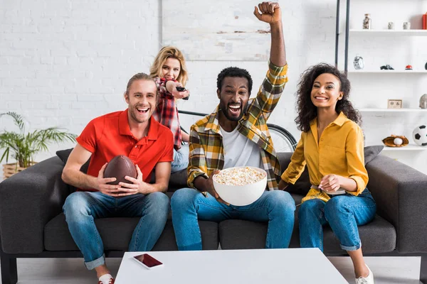 Happy multicultural friends sitting on couch and watching championship near table with popcorn in bowl — Stock Photo