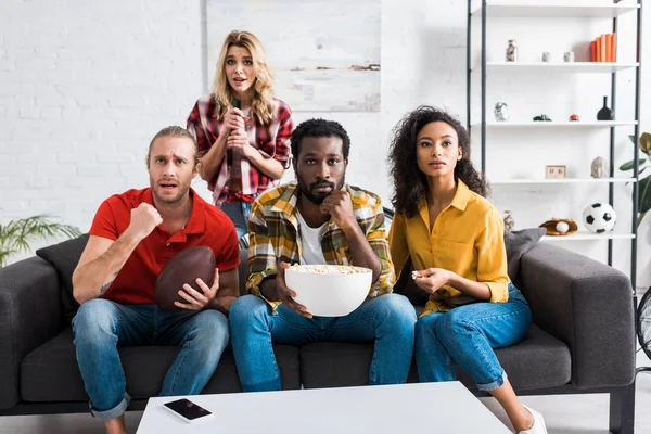 Sorpresi e sconvolti amici multiculturali seduti sul divano e guardando la partita vicino al tavolo in soggiorno — Foto stock