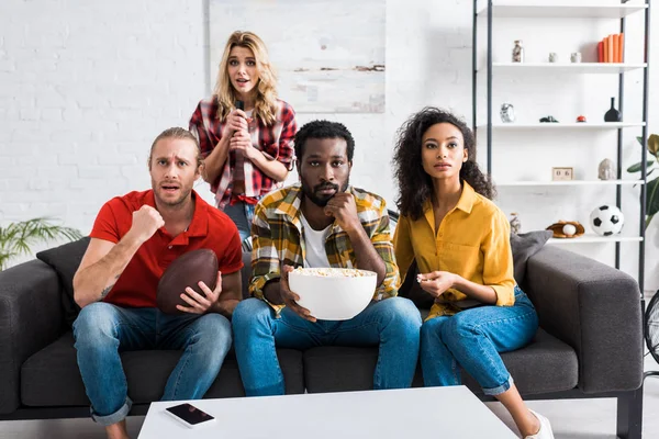 Surpreso e chateado amigos multiculturais sentados no sofá e assistindo jogo perto da mesa na sala de estar — Fotografia de Stock