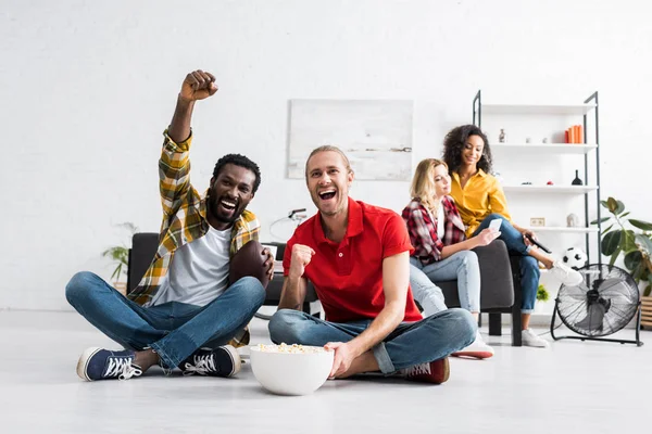 Jóvenes multiculturales felices y positivos sentados en el suelo y viendo un partido deportivo con palomitas de maíz - foto de stock