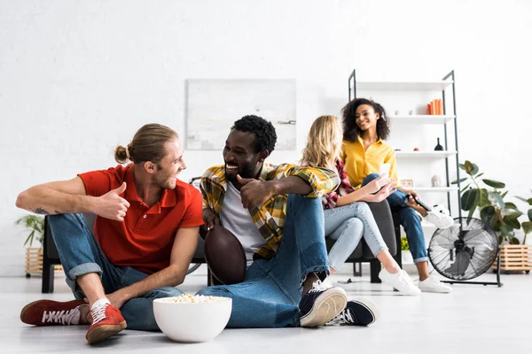 Выборочный фокус двух улыбающихся мультикультурных мужчин, сидящих на полу и разговаривающих с миской попкорна — стоковое фото