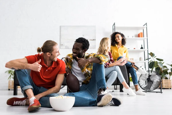 Selective focus of two smiling multicultural men sitting on floor and talking with bowl of popcorn — Stock Photo