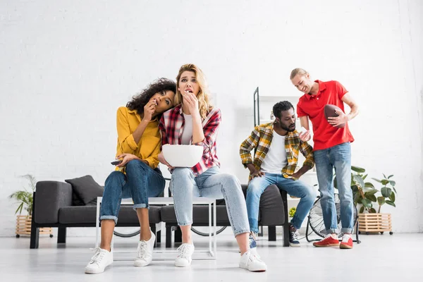 Enfoque selectivo de dos mujeres multiculturales viendo partido y comiendo sabrosas palomitas de maíz en la sala de estar - foto de stock