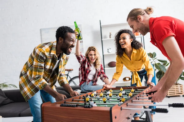 Cuatro felices y alegres amigos multiétnicos jugando futbolín en la sala de estar - foto de stock