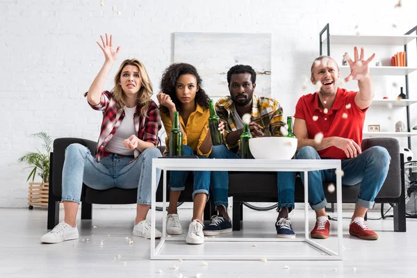 Cuatro amigos multiétnicos disgustados sentados en el sofá, viendo películas y lanzando palomitas de maíz - foto de stock