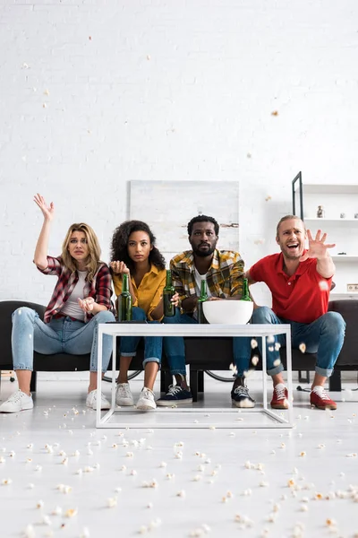 Selective focus of four disappointed multicultural friends sitting on couch near table with bowl of popcorn and watching movie — Stock Photo