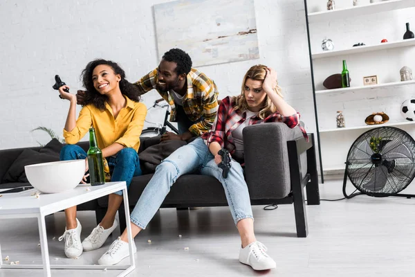 Vista de ángulo bajo de chica molesta sentada cerca de amigos afroamericanos felices en la sala de estar - foto de stock