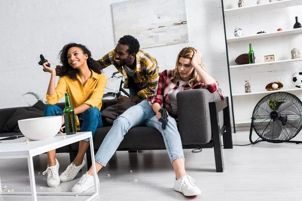 Vue à angle bas de fille bouleversée assise près d'amis afro-américains heureux dans le salon — Photo de stock