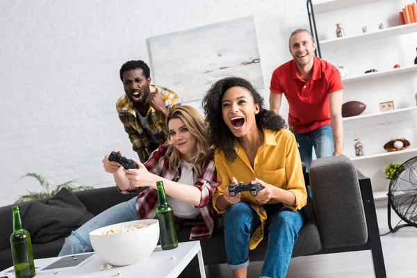 Chicas multiculturales emocionadas jugando videojuego cerca de los hombres en la sala de estar - foto de stock