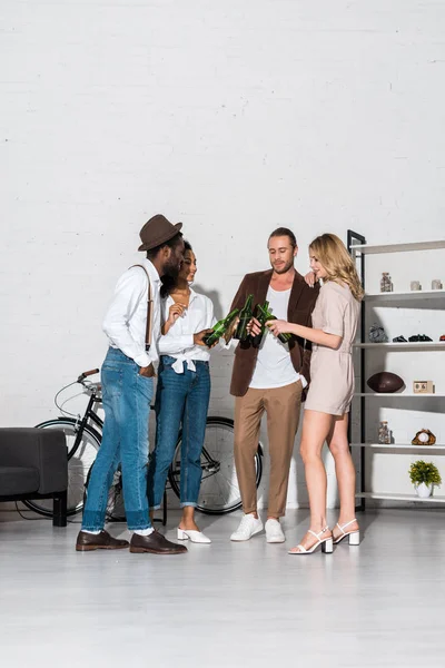 Happy stylish multicultural friends clinking bottles of beer at home — Stock Photo