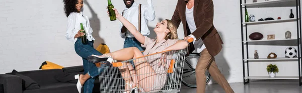 Panoramic shot of happy stylish girl riding in shopping cart near multicultural friends holding bottles of beer — Stock Photo