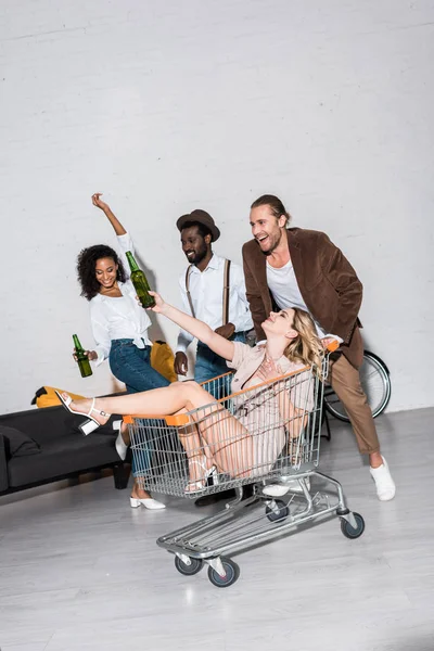 Chica alegre montar en el carrito de compras cerca de amigos felices multiculturales — Stock Photo