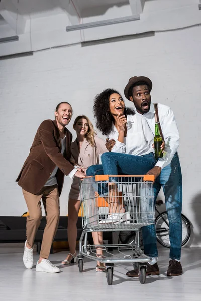 Visão de baixo ângulo da menina americana africana feliz montando no carrinho de compras perto de amigos multiculturais — Fotografia de Stock