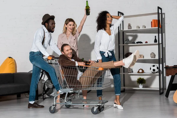 Hombre alegre cabalgando en la tarjeta de compras cerca de amigos multiculturales felices - foto de stock
