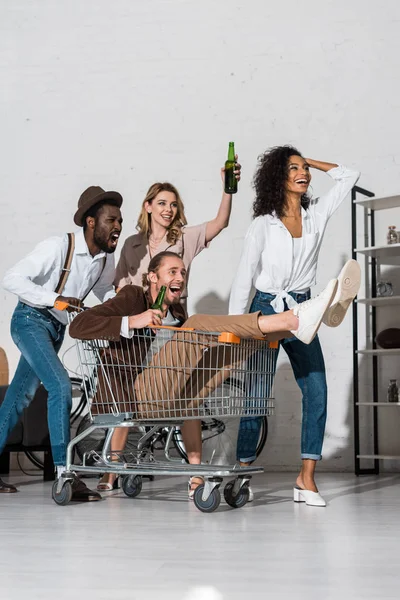 Fröhlicher Mann reitet in Einkaufskarte und hält Flasche in der Nähe fröhlicher multikultureller Freunde — Stockfoto