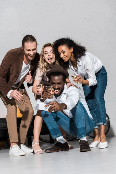 Vista basso angolo di felice uomo afro-americano prendendo selfie con gli amici in possesso di bicchieri di champagne — Foto stock