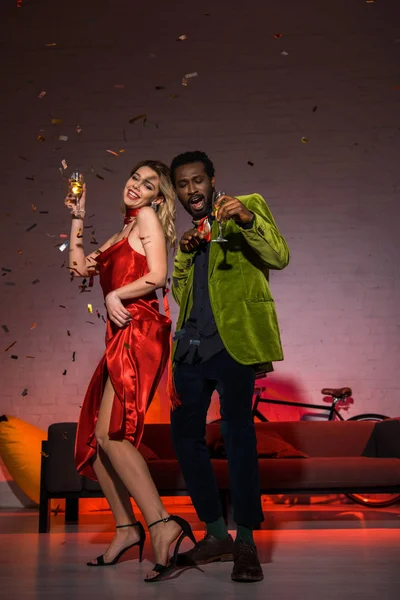 Vista de ángulo bajo de hombre afroamericano feliz de pie con mujer rubia en vestido rojo y celebración de copa de champán - foto de stock