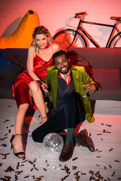 Junge blonde Frau in rotem Kleid sitzt neben afrikanisch-amerikanischem Mann mit Champagnerglas — Stockfoto