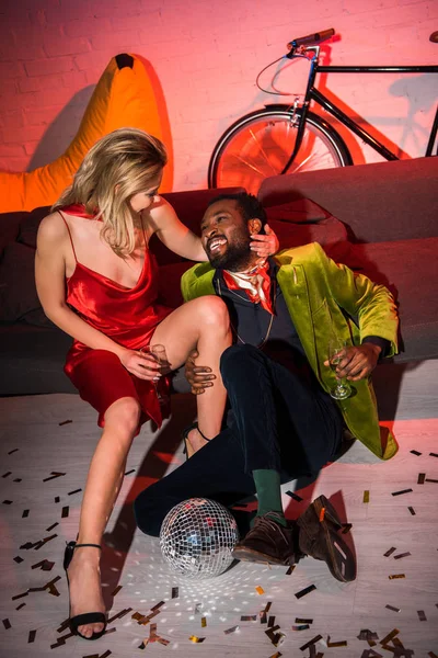 Joven rubia en vestido rojo sentado con hombre afroamericano en club nocturno - foto de stock