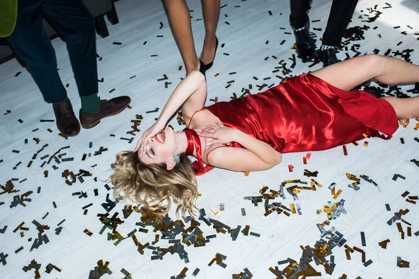 Menina loira atraente e bêbada em vestido vermelho deitado no chão com confete perto de amigos — Fotografia de Stock