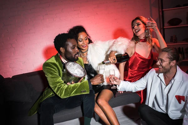 African american woman holding bottle near champagne glass while sitting near multicultural friends — Stock Photo