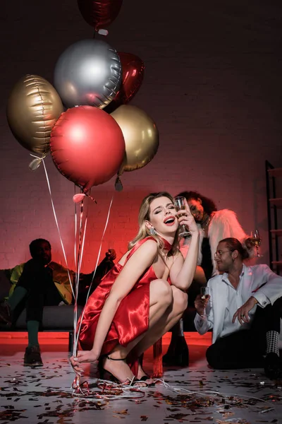 Вибірковий фокус п'яної блондинки в червоній сукні, що тримає повітряні кулі та келих шампанського біля мультикультурних друзів — стокове фото