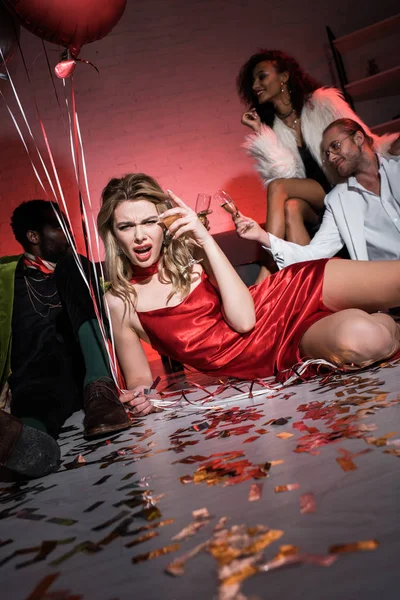 Selective focus of tired girl in red dress lying on floor and holding balloons near multicultural friends — Stock Photo