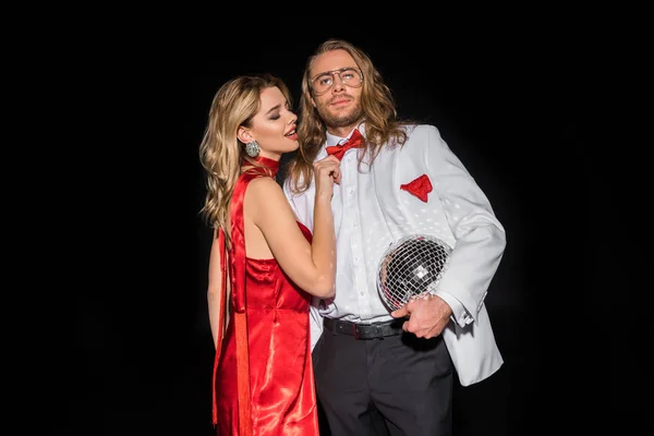 Attractive woman in red dress touching bow tie of man in glasses with disco ball isolated on black — Stock Photo