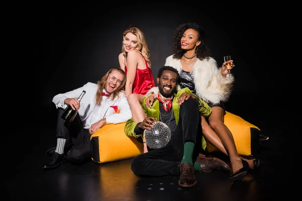 Happy african american man holding dicso ball while sitting near bean bag chair and friends  on black — Stock Photo