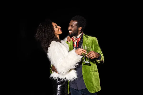 Homem americano africano alegre olhando para a mulher americana africana segurando copo de champanhe e sorrindo isolado no preto — Fotografia de Stock