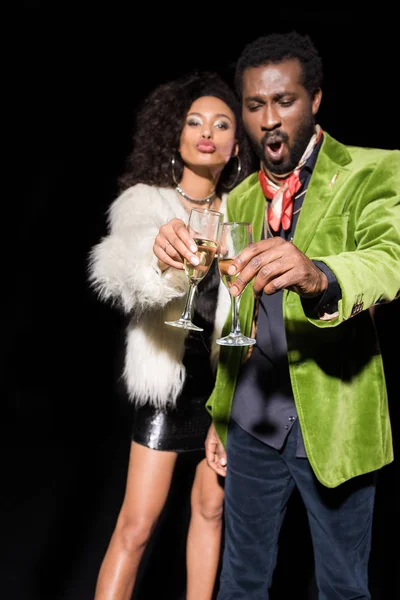 Selektiver Fokus eines überraschten afrikanisch-amerikanischen Mannes und einer Frau mit Entengesicht, die Champagnergläser auf schwarzem Grund klirren — Stockfoto