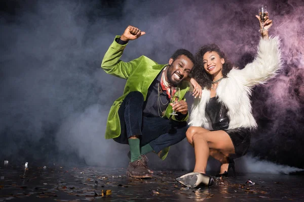 Happy african american man and woman holding champagne glasses while sitting near confetti on black with smoke — Stock Photo