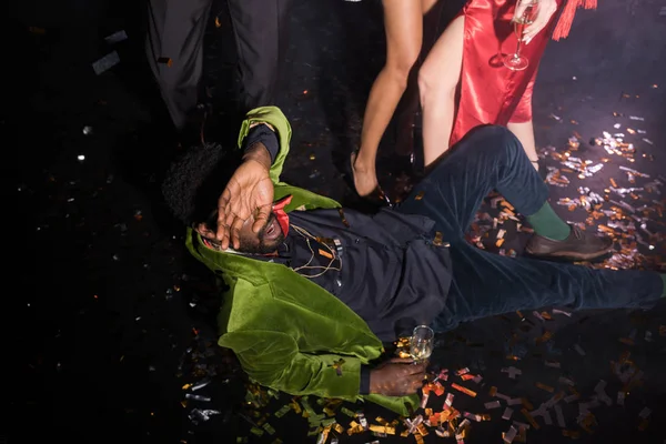 Drunk african american man covering face while lying on floor with confetti near friends on black — Stock Photo