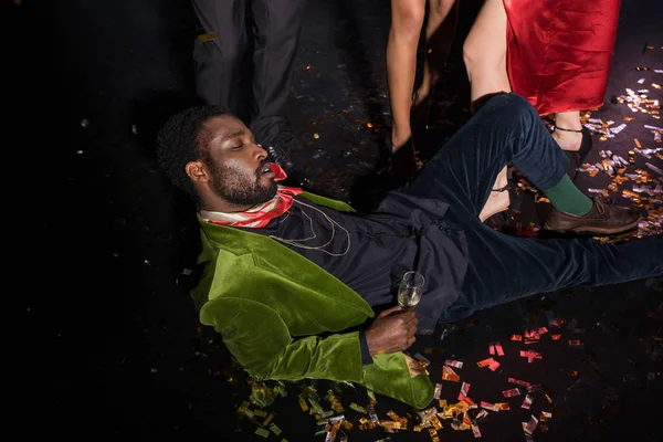 Bonito e bêbado afro-americano homem deitado no chão com confete perto amigos no preto — Fotografia de Stock