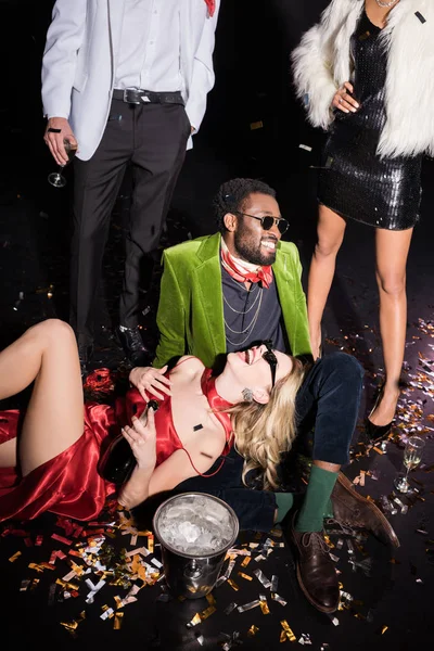 Happy woman in sunglasses lying near african american man and friends on black — Stock Photo