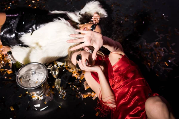 Overhead view of blonde girl in red dress gesturing while smiling and lying on floor with confetti near african american friend on black — Stock Photo