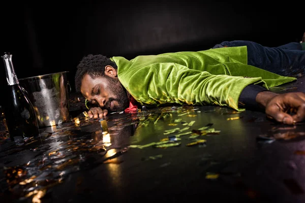 Messa a fuoco selettiva di ubriaco uomo afroamericano sdraiato sul pavimento con coriandoli vicino secchio di ghiaccio e bottiglia di champagne sul nero — Foto stock