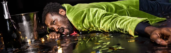 Plan panoramique de l'homme afro-américain ivre couché sur le sol avec des confettis brillants près du seau à glace et bouteille de champagne sur noir — Photo de stock