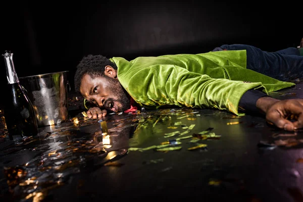 Selektiver Fokus eines betrunkenen afrikanisch-amerikanischen Mannes auf dem Boden liegend mit glänzendem Konfetti in der Nähe von Eiskübeln und einer Flasche Champagner auf schwarz — Stockfoto