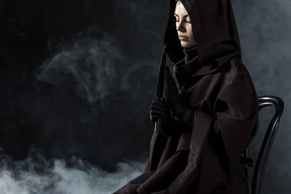Mujer en traje de la muerte sosteniendo cuchillo y sentado en silla en negro - foto de stock