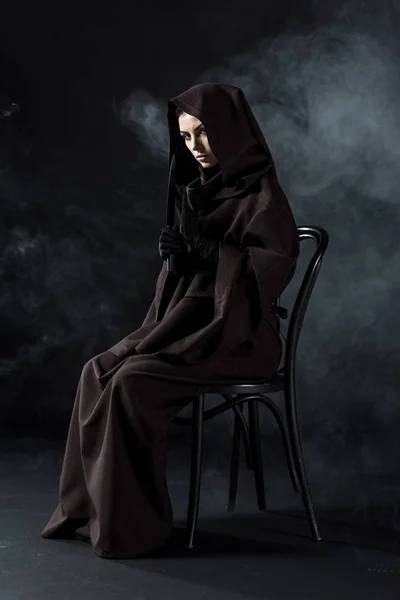 Женщина в костюме смерти держит нож и сидит на стуле на черном — стоковое фото