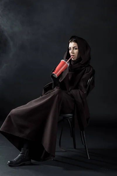 Frau im Todeskostüm sitzt auf Stuhl und trinkt Getränk auf schwarz — Stockfoto