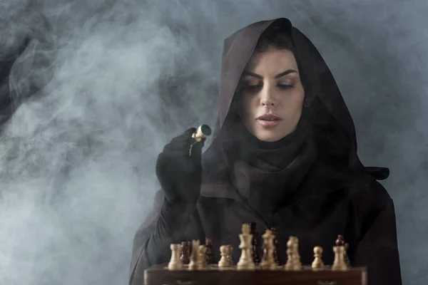 Woman in death costume playing chess in smoke on black — Stock Photo