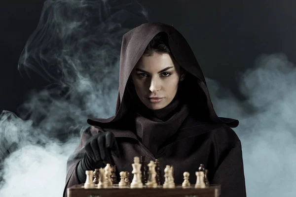 Woman in death costume playing chess in smoke on black — Stock Photo