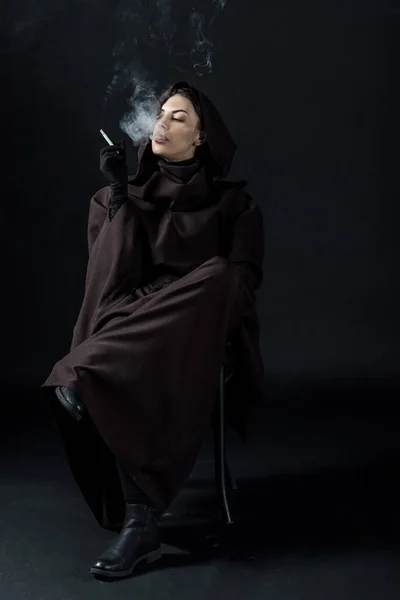 Nachdenkliche Frau im Todeskostüm sitzt auf Stuhl und raucht Zigarette auf Schwarz — Stockfoto